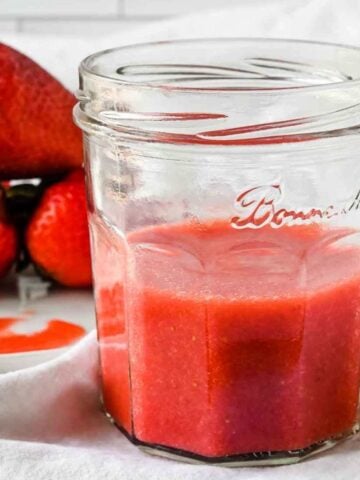 close up shot of strawberry puree in a jar with strawberries behind on a white background.