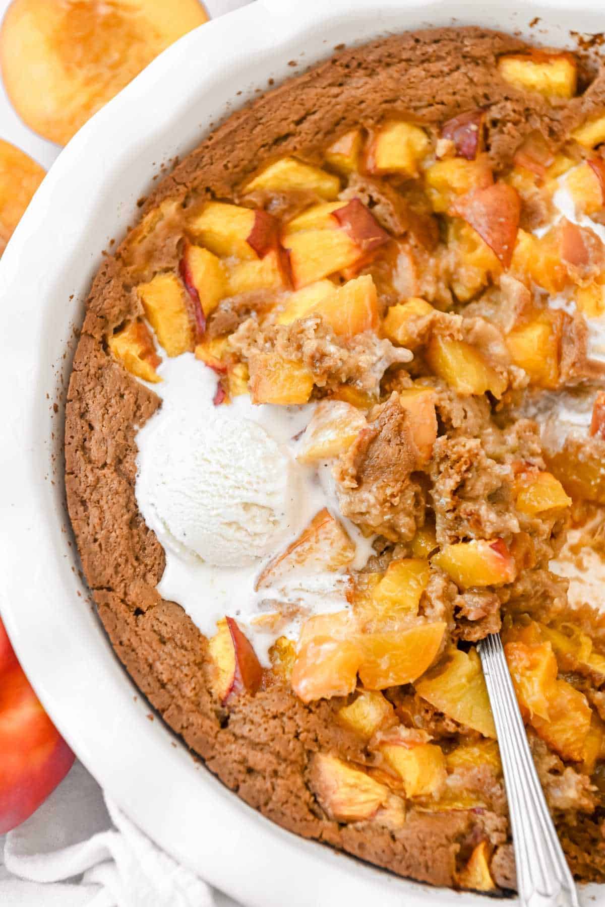 close up of nectarine cobbler in a white pie plate with a spoon, topped with vanilla ice cream.