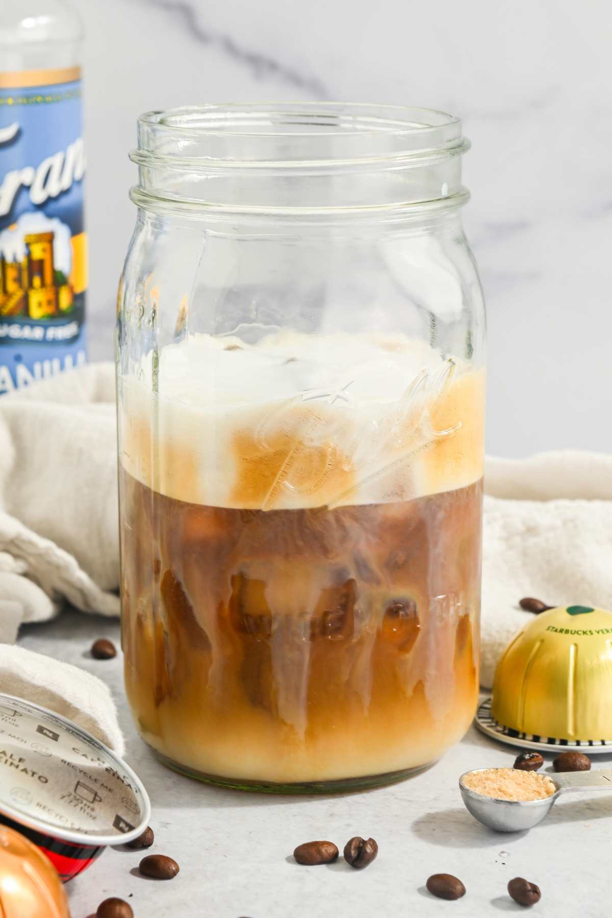 Nespresso coffee in a mason jar with foam on top and with capsules and coffee beans around it.